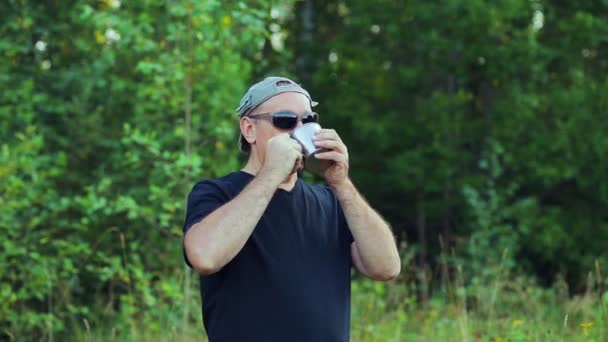 En man på kanten av skogen dricker te från en mugg och beundrar naturen. — Stockvideo