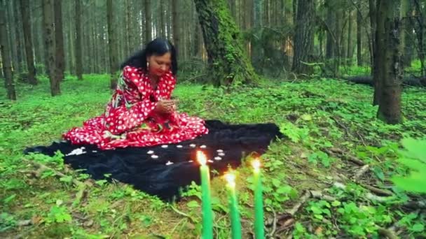 Een jonge zigeunervrouw in het bos gooit kiezels en magische bewegingen maakt met haar handen over het branden van kaarsen. — Stockvideo