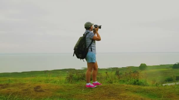 A woman with a backpack behind her back on the mountain takes a picture of the lake. — Stock Video
