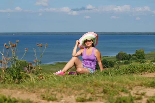 Joven mujer feliz en un sombrero y gafas de sol se sienta en el borde de una colina frente a un lago . —  Fotos de Stock