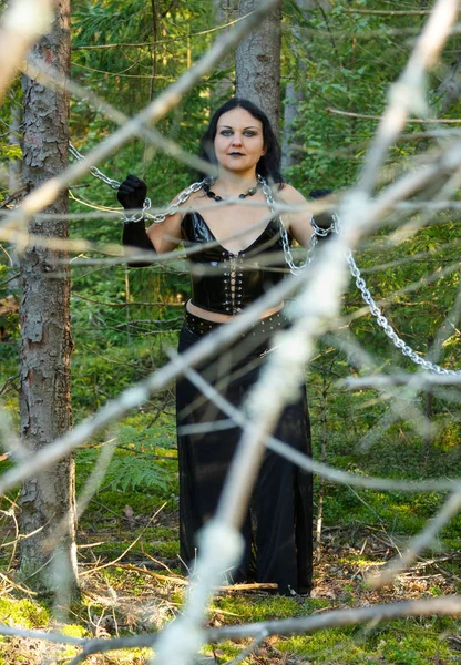 Eine Frau ist eine Hexe in Schwarz in Ketten zwischen Bäumen in einem dichten Wald. halloween. — Stockfoto