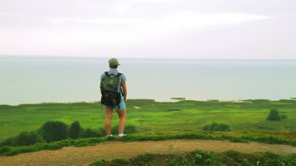 Reisenden mit einem Rucksack auf dem Rücken steht am Rande des Hügels und bewundert den See im Regen. Gesamtplan. — Stockvideo