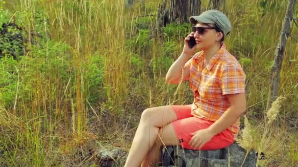 Mladá žena turista v sluneční brýle a baseballovou čepici v lese sedí na padlý strom a mluví o telefonu. — Stock video