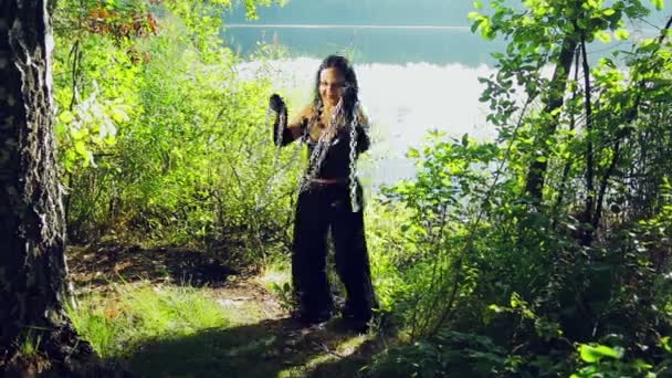 Una mujer riendo de una bruja vestida de negro en la orilla del lago se está divirtiendo y bailando con cadenas en las manos. Halloween. Estilo Gothick . — Vídeo de stock