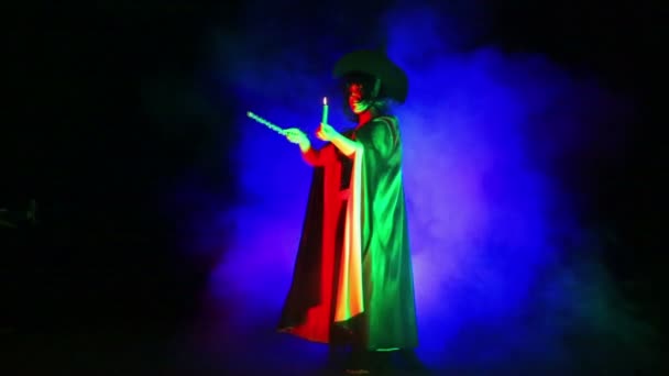 Молода відьма в чорному плащі і капелюсі зі свічкою в руці в хмарі диму займається магією — стокове відео