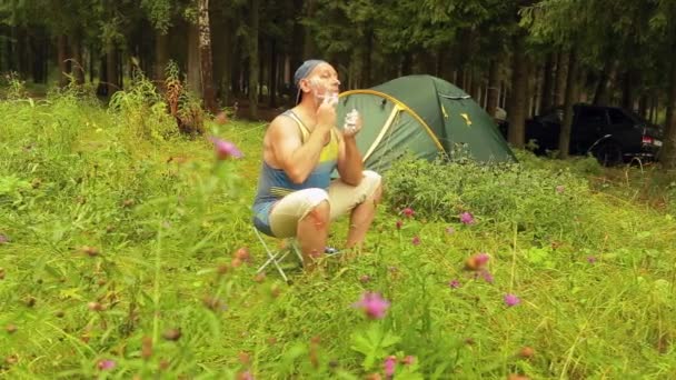 Een toeristische man zit in de buurt van een tent en scheert zijn gezicht. — Stockvideo