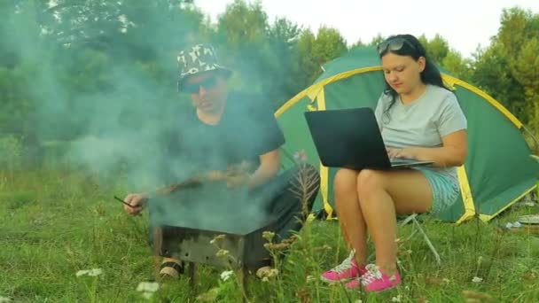 Een man en een vrouw zijn in de buurt van een tent. Een vrouw die werkt op een laptop, draait een man om een grill met vlees. — Stockvideo