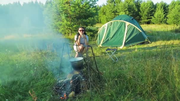 Mladá žena sedí u ohně u stanu, pije čaj a sleduje tvrďák, visí nad ohněm. — Stock video