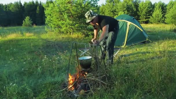 Ein Mann mit einem Becher Tee in der Hand schiebt Holz in das Feuer, über dem die Melone hängt. — Stockvideo