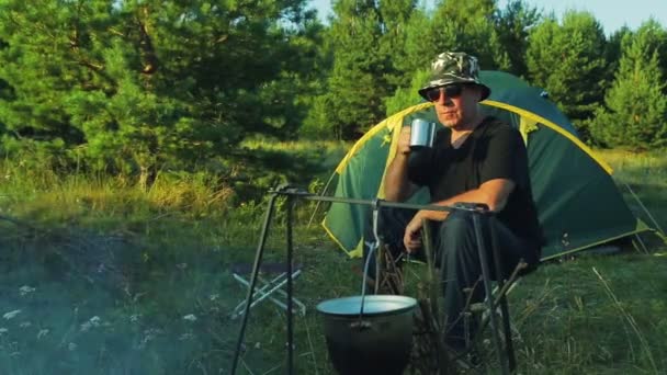 Een man in zonnebril zitten in de buurt van een bowler opknoping boven een vuur in de buurt van een tent, het drinken van thee en het bewonderen van de natuur. — Stockvideo