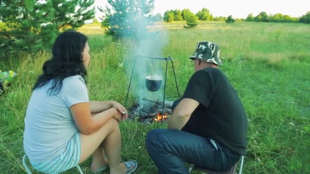 Egy férfi és egy nő ül a tűz, amely felett a Dobó lóg. az ember moccan a parazsat a tűz. — Stock videók