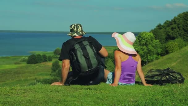 Pár muž a žena s batohy sedí na okraji hory a obdivovat výhled na jezero. Střelba ze zadní. — Stock video