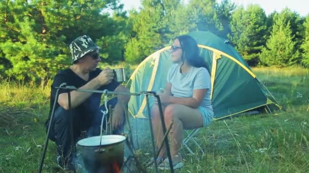 Man och kvinna turister sitter på tältet, dricka te och titta på potten hängande över elden. — Stockvideo