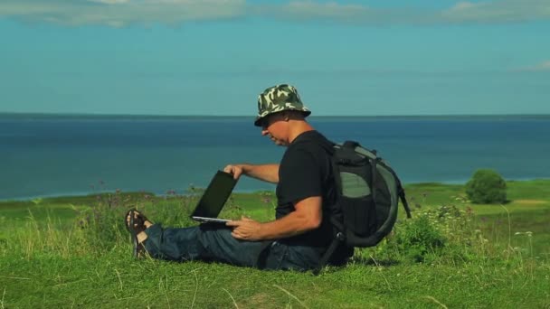 Ein Mann mit einem Rucksack auf dem Rücken sitzt am Rande des Berges und öffnet einen Laptop. Schuss von der Seite. — Stockvideo