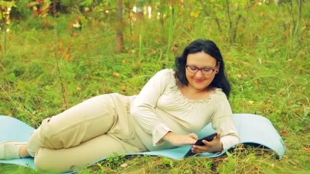 A young smiling woman in glasses in the forest lies on a rug for yoga and communicates in a messenger. — Stock Video