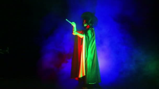 Una joven bruja con una capa negra y un sombrero con una vela en la mano en una nube de humo se dedica a la magia — Vídeo de stock