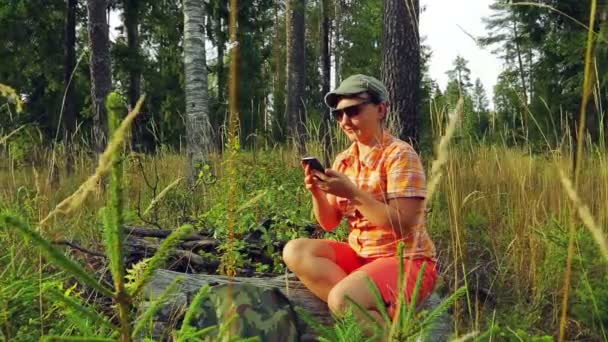 Молода жінка турист в сонцезахисних окулярах і бейсбольна шапка в лісі в сонячний день сидить на падіння дерева і спілкується по телефону в посланні . — стокове відео