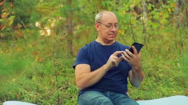Чоловік в окулярах в лісі сидить на траві і спілкується в посланні на планшеті . — стокове відео
