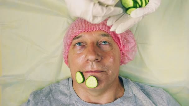 Las manos de la mujer del cosmético ponen a la persona del hombre la máscara de los trozos del pepino . — Vídeo de stock