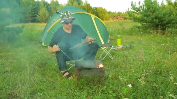 Un uomo siede vicino alla tenda e griglia la carne sulla griglia e mescola i carboni . — Video Stock