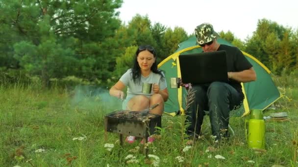 Un uomo e una donna vicino alla tenda nella natura friggono shish kebab e bevono tè. Un uomo sta lavorando su un portatile . — Video Stock