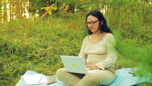 Una mujer morena con gafas en un claro del bosque trabajando con un portátil . — Vídeos de Stock