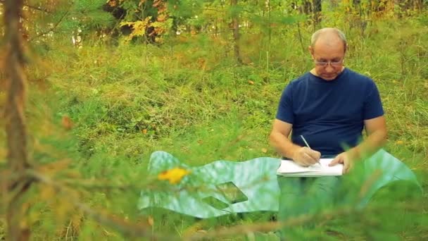 Een man in bril aan de rand van het bos werkt met schetsen in potlood in een notitieblok. — Stockvideo