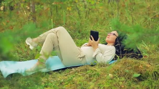 Молода жінка в окулярах лежить на траві в парку і спілкується в посланні на планшеті . — стокове відео