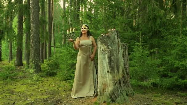 A forest fairy in a golden mask on his face stands at a large stump and holds a candlestick with candles in his hand. — Stock Video