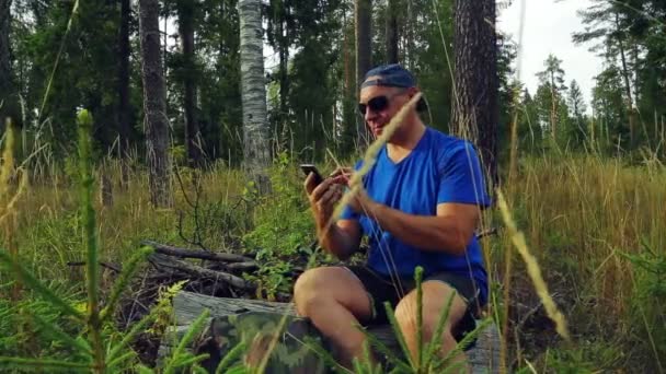 一个在森林里的太阳镜和棒球帽的游客坐在一棵倒下的树上 在一个信使的电话里交流 — 图库视频影像