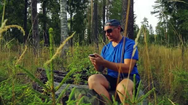Турист в сонцезахисних окулярах і бейсбольна шапка в лісі на сонячний день сидить на падаючому дереві і спілкується по телефону в посланні . — стокове відео