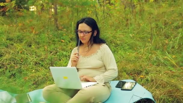 Una mujer pensativa en gafas en el borde del bosque con un lápiz en la mano y una computadora portátil encuentra una solución . — Vídeo de stock