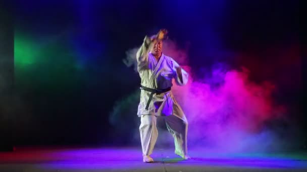 Un hombre se dedica al karate sobre un fondo negro con humo de color — Vídeo de stock