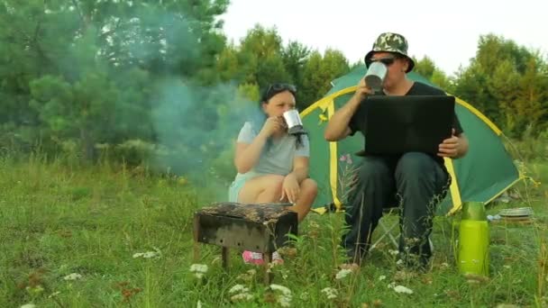 Een man en een vrouw in de buurt van de tent barbecue vlees op de grill, drinken thee en werken op een laptop. — Stockvideo