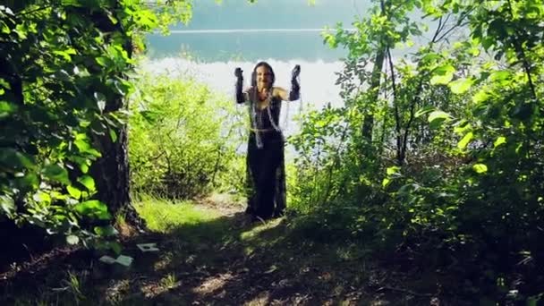 Een lachende vrouw van een heks in zwarte kleren aan de oever van het meer is plezier en dans met ketens in haar handen. Halloween. Gothick stijl. — Stockvideo