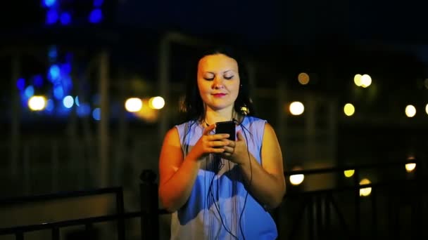 Молодая улыбающаяся женщина на набережной в наушниках слушает музыку с телефона и поет на фоне ночного неба — стоковое видео