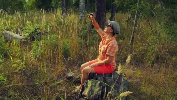 Girl tourist in sunglasses and a baseball cap in the forest sits on a fallen tree and takes pictures on the phone. — Stock Video