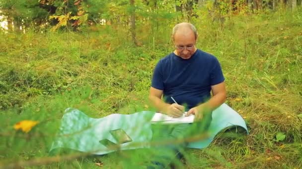 En man i glasögon vid kanten av skogen ritar diagram i en penna med en penna. — Stockvideo