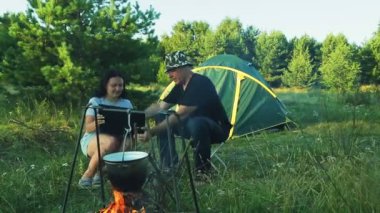 Erkek ve kadın turist çadırı, ateşin üzerinde asılı ve bir dizüstü bilgisayar ile çalışma potu izliyor oturuyorlar.
