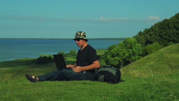 Людина з рюкзаком, що сидить на краю гори з видом на озеро і працює з ноутбуком. закриває ноутбук і видаляє його . — стокове відео