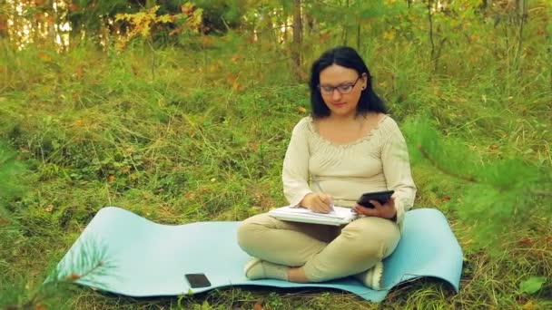 Uma jovem mulher em óculos em uma clareira da floresta trabalhando em um tablet e lápis em um caderno — Vídeo de Stock