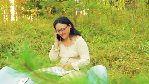 Uma jovem mulher na floresta está trabalhando com um lápis em um caderno e falando em seu celular — Vídeo de Stock