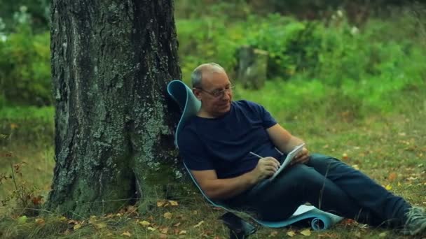 Ένας χαμογελαστός άνθρωπος γυαλιά κάθεται κάτω από ένα δέντρο στο πάρκο και να γράφει σε ένα σημειωματάριο με ένα μολύβι. — Αρχείο Βίντεο