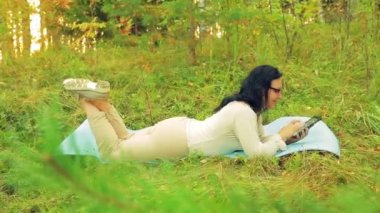 Bir tablet ile yoga ve çalışmaları için bir halı üzerinde bir orman içinde genç bir kadın yatıyor.