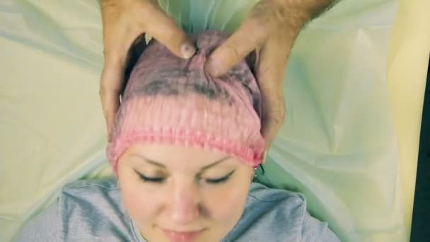Руки чоловіка косметолога одягнені на голову жінки, що лежить на дивані косметична шапка . — стокове відео