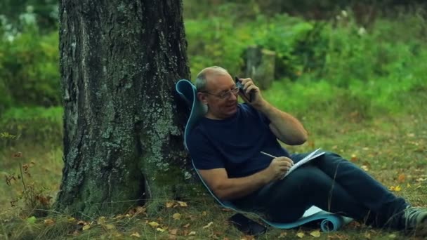 Ένας χαμογελαστός άνθρωπος γυαλιά κάθεται κάτω από ένα δέντρο στο πάρκο και αντλεί ένα σημειωματάριο, μολύβι και συνομιλίες στο τηλέφωνο — Αρχείο Βίντεο