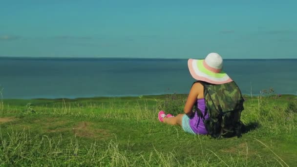 Una mujer en un sombrero de paja con una mochila detrás de su espalda se sienta en una montaña y admira la naturaleza. disparando por la espalda . — Vídeos de Stock