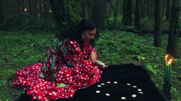 Zigeuner shuvani in het bos in de schemering maakt magische bewegingen met handen over het branden van kaarsen en witte kiezels. — Stockvideo