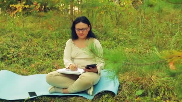 Μια νεαρή γυναίκα σε ποτήρια στην άκρη του δάσους του λειτουργεί σε ένα δισκίο και αντλεί ένα μολύβι σε ένα σημειωματάριο — Αρχείο Βίντεο
