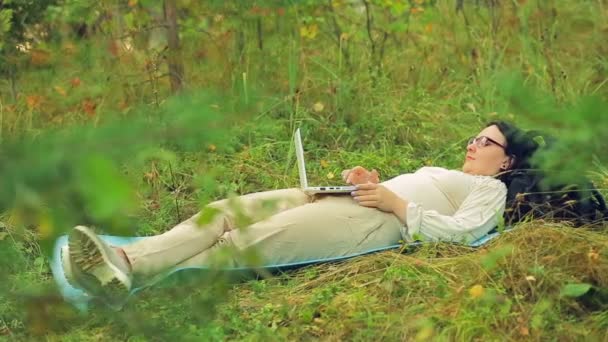 Молода жінка в окулярах лежить на траві в парку і працює з ноутбуком — стокове відео